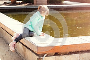 Lonely little caucasian girl sitting by the lake water and looking at the water