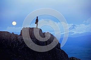 Lonely man at evening stands on cliff against mountains and moon