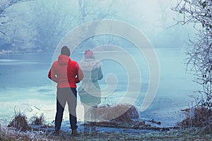 Lonely man in red jacket standing by the lake in winter, with transparent woman figure standing next to him