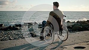 Lonely man is sitting in invalid carriage on sea shore, looking in distance