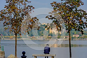 Lonely man sitting between two trees on a bench and looking at the lake. In the distance you can see the island on which stands th