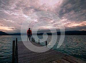 Lonely man standing on the edge of the wooden pier looking at the calm lake and forest on the other side