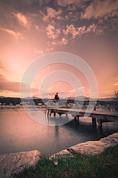 Lonely man standing on the edge of the wooden pier looking at the calm lake and forest on the other side