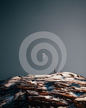 Lonely man on the top of a snow-capped mountain admiring the daytime half-moon during sunset