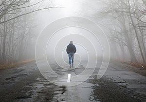 Lonely man walking in fog away road. Rural landscape with road in morning mist. Warm autumn colors. Dark mysterious background