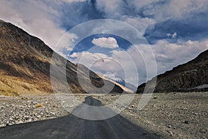 A Lonely Metal Road amidst Ladakh\'s Empty Valley, with Clear Blue Sky, Fluffy Clouds, and Majestic Snowy Mountains in Sight.