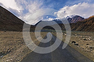 A Lonely Metal Road amidst Ladakh\'s Empty Valley, with Clear Blue Sky, Fluffy Clouds, and Majestic Snowy Mountains in Sight.