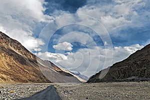 A Lonely Metal Road amidst Ladakh\'s Empty Valley, with Clear Blue Sky, Fluffy Clouds, and Majestic Snowy Mountains in Sight.