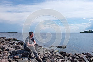 A lonely middle-aged man sits on a rocky beach on a summer day and looks thoughtfully into the distance.