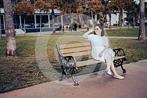 Lonely middle-aged woman sits alone on a bench in a park area on the waterfront. wearing a white dress and sunglasses. an adult