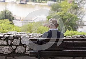 A lonely old man sitting on a bench in a park, looking at river