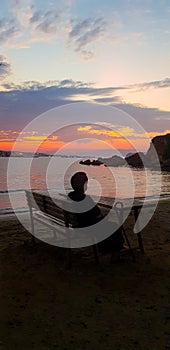 Lonely old woman in the evening sitting on a bench on the beach and watching the beautiful sunset