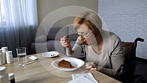 Lonely retired lady in wheelchair eating slowly her lunch at the table, hospital