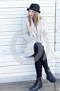 Lonely sad beautiful girl in a black coat and hat, sitting on a white bench cold winter sunny day