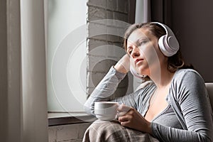 Lonely sad beautiful woman with cup of coffee listenning music in headphones sitting on sofa near window at morning.