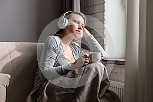 Lonely sad beautiful woman with cup of coffee listenning music in headphones sitting on sofa near window at morning.