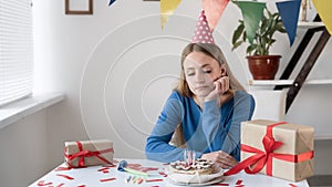 A lonely sad girl business woman sits at a table with a cake and gifts with her head in a cap on her hand. A bored young