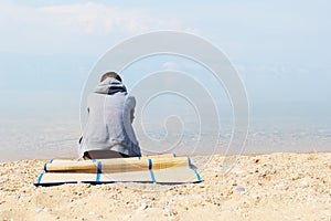 A lonely sad girl is sitting on the beach in a gray hoodie. Back view