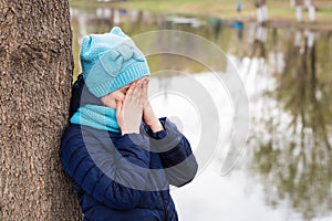 A lonely sad girl stands covering her face with the hands of a tree on the shore of the lake. Mental health. Teenage years