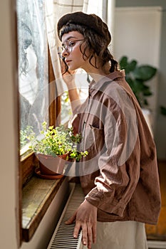 Lonely sad hipster girl looking out window while spending time at home