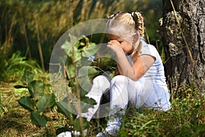 Lonely sad little girl in a white dress and a flower in her hand was lost in the woods, sitting near a tree and crying during day