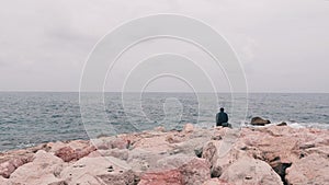 Lonely sad man sitting on big stone near ocean. Office worker sitting near sea after work day. Isolated lonely boy looking at sea.