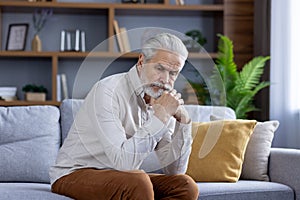 A lonely senior gray-haired man sits upset at home on the sofa, and pensively contemplates, resting his head on two