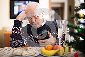 lonely senior man sitting at home table during Christmas holidays, looking at phone