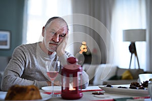 Lonely senior man with wine sitting at the table indoors at Christmas, solitude concept.