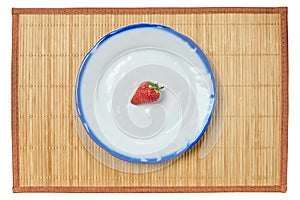 Lonely strawberry on a white plate with a blue rim on a cane place mat