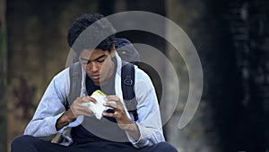 Lonely teenager holding sandwich sitting outdoors, having snack, solitude