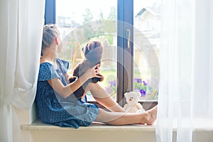 Lonely toddler russian girl sitting near window at home playing