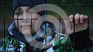 Lonely unhappy, miserable young boy behind and clinging to fence