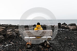 Lonely unrecognized traveler man in yellow rain jacket  is relaxing at nice view of sea with wooden bench in Iceland