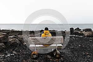 Lonely unrecognized traveler man in yellow rain jacket  is relaxing at nice view of sea with wooden bench in Iceland photo