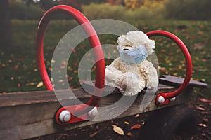 Lonely white teddy bear soft toy with disposal face mask sits on a wooden outdoor seesaw in times of social distancing