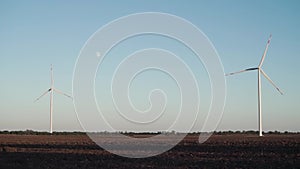 a lonely wind turbine stands in an unseeded field. the moon is visible in the sky, summer sunset