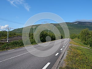 Lonely winding asphalt road and railroad in Abisko village and national park with birch forest and mountains, summer blue sky