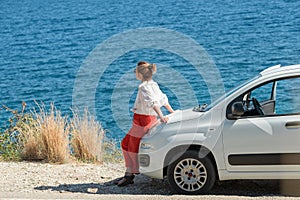 Lonely woman dressed in light summer clothes near the parked on roadside little white car enjoying the Ionian sea landscape on