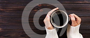 Lonely woman drinking coffee in the morning, top view of female hands holding cup of hot beverage on wooden desk. Banner