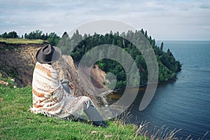 Lonely woman in felt hat and woolen poncho sitting on grassy seashore and looking at wooded cape. Travel, adventure, feeling