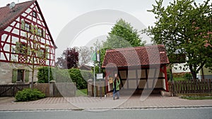 Lonely woman in green coat and backpack waiting for a bus near Fachwerk style bus stop, Bavaria, Germany, general plan