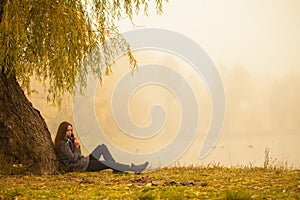 Lonely woman having rest under the tree near the water in a foggy autumn day