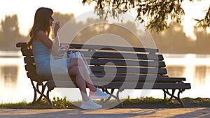 Lonely woman sitting alone on lake shore bench on warm summer evening. Solitude and relaxing in nature concept