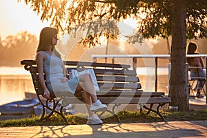 Lonely woman sitting alone on lake shore bench on warm summer evening. Solitude and relaxing in nature concept