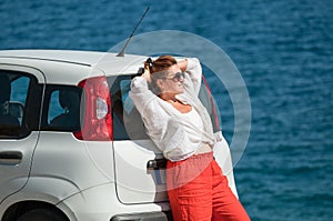 Lonely woman in sunglasses dressed light summer clothes near parked on roadside little white car enjoying Ionian sea landscape on