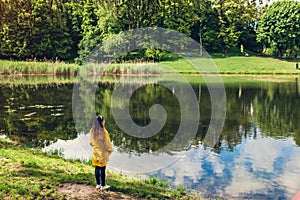 Lonely woman in yellow raincoat walking in summer park. Girl admires lake landscape view after rain