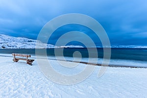 Lonely wooden bench overlooking the sea on a snow covered beach at dusk