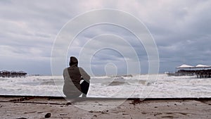 Lonely young caucasian man sitting on shore of stormy winter sea. Man pull on hood on head. Back view