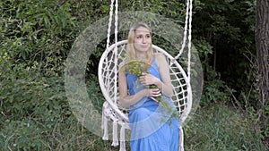A lonely young girl with a bouquet of yellow flowers swinging on a white hanging swing in a blue dress at the sunset of